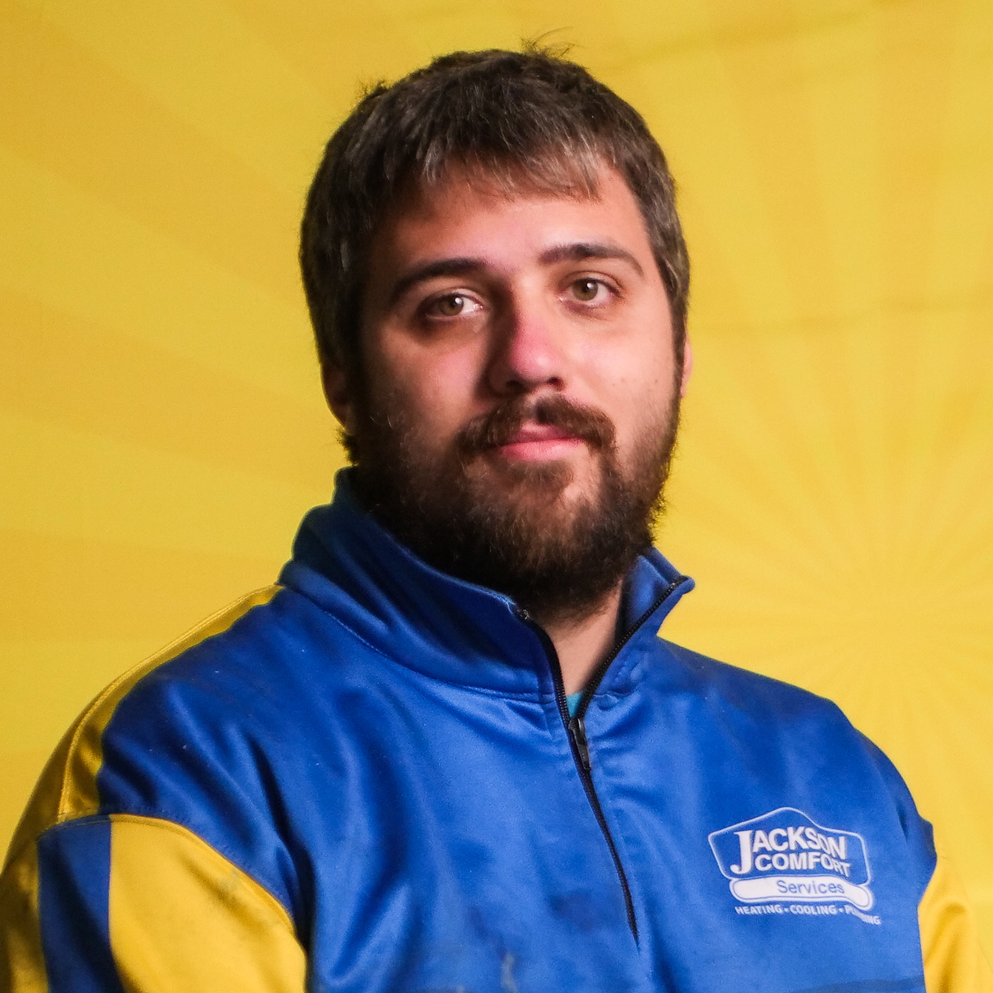 A Man With Dark Hair And A Beard, Representing Our Team, Wears A Blue And Yellow Jackson Comfort Services Jacket As He Stands Against A Vibrant Yellow Background.