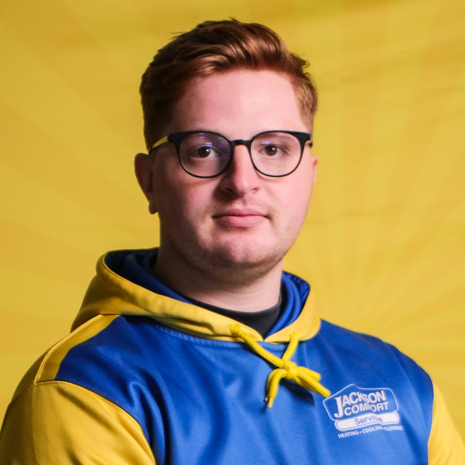 A Member Of Our Team With Short Red Hair And Glasses Wears A Blue And Yellow &Quot;Jackson Comfort Services&Quot; Hoodie, Posed In Front Of A Yellow Background.