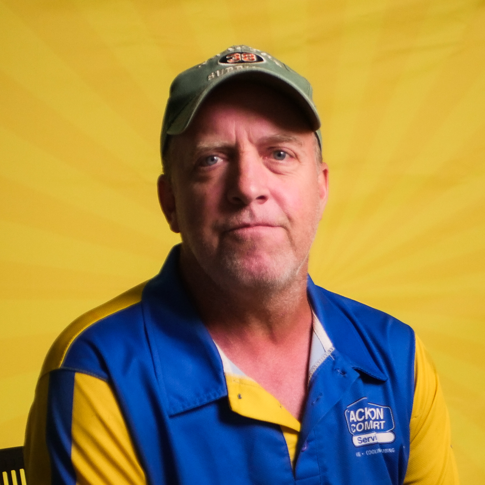 A Member Of Our Team, Wearing A Green Cap And A Blue And Yellow Action Comfort Services Shirt, Sits In Front Of A Yellow Background With A Sunburst Pattern.