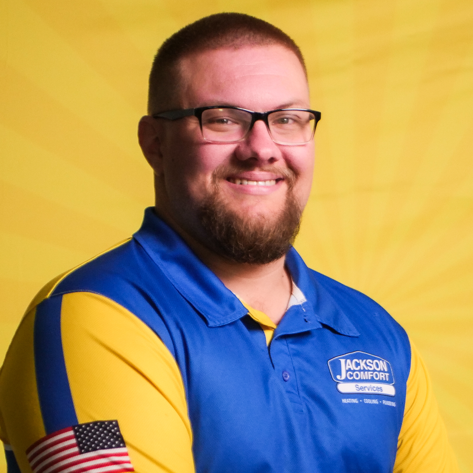 A Member Of Our Team With Glasses And A Beard Smiles At The Camera, Wearing A Blue And Yellow Jackson Comfort Services Shirt With A U.s. Flag Patch On The Sleeve, Set Against A Yellow Background.
