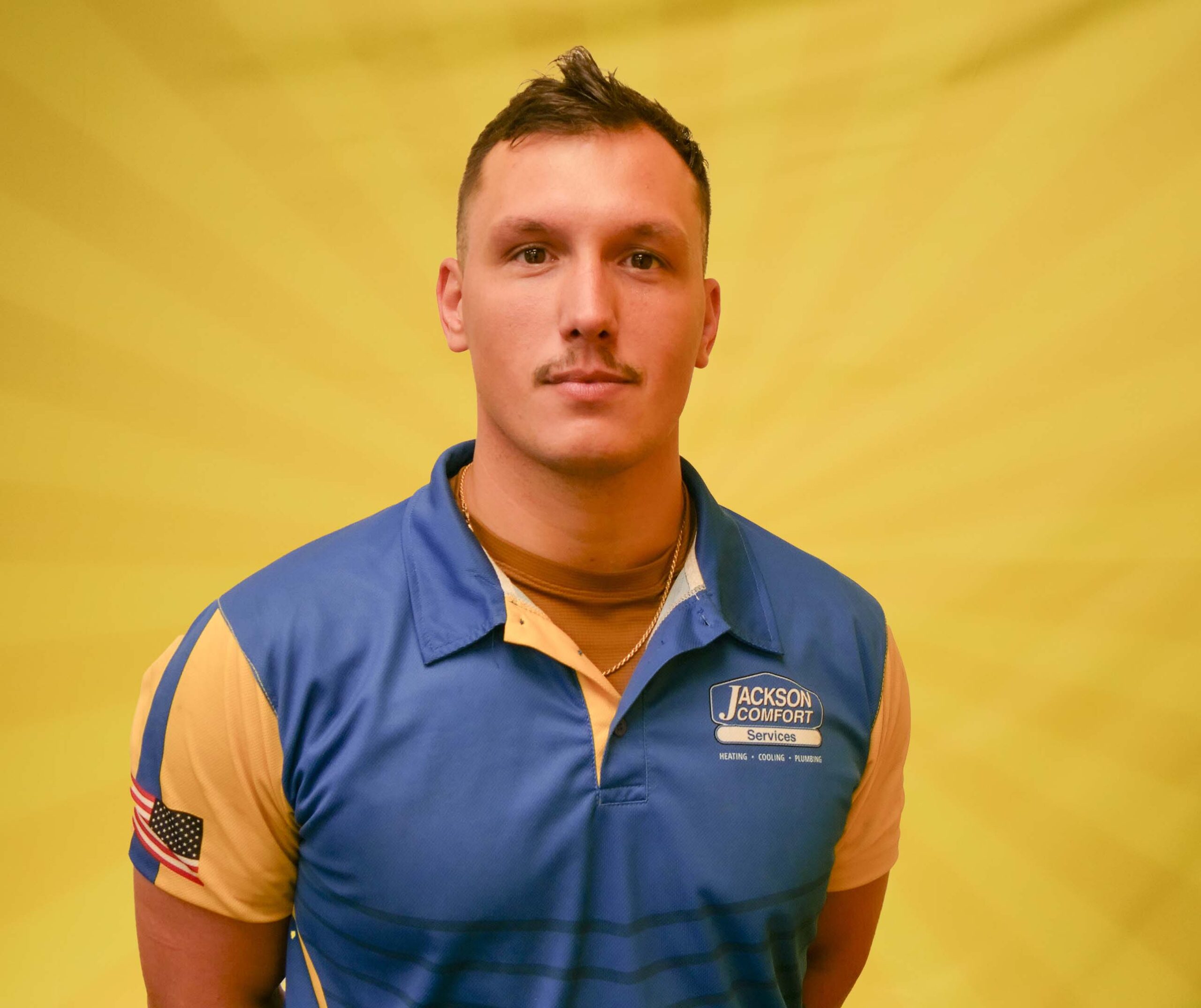 A Member Of Our Team, Wearing A Blue And Yellow Jackson Comfort Services Shirt, Stands In Front Of A Yellow Background.