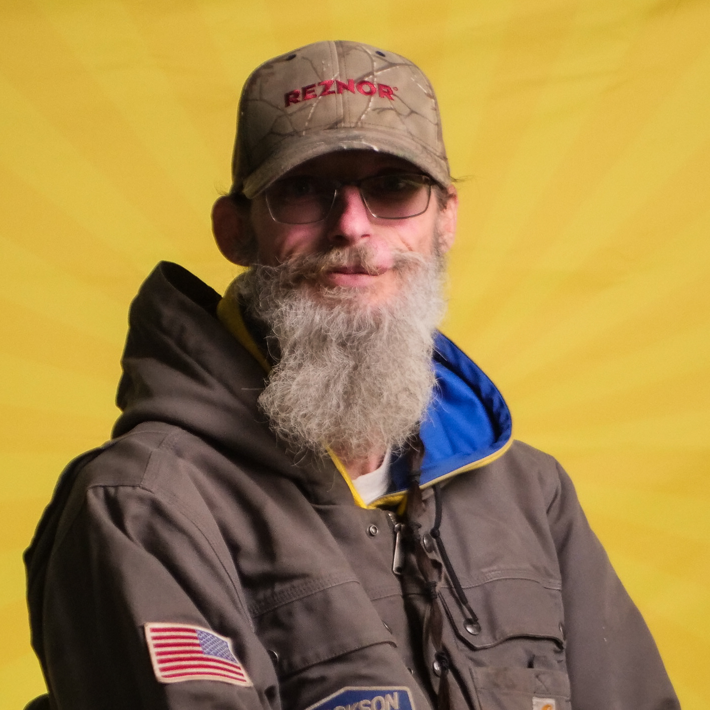 A Member Of Our Team With A Gray Beard And Glasses, Wearing A Tan Cap, Brown Jacket, And An American Flag Patch, Poses In Front Of A Yellow Background.
