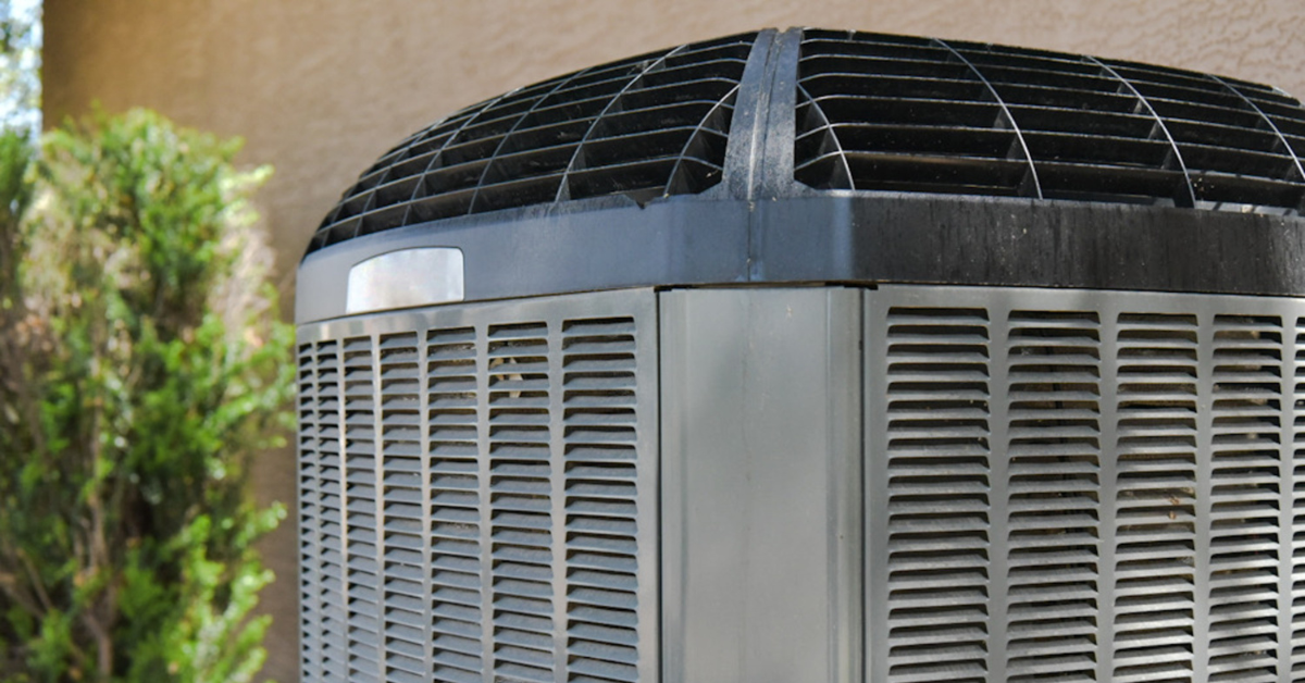 Outdoor central air conditioning unit next to a building, surrounded by plants and receiving sunlight.