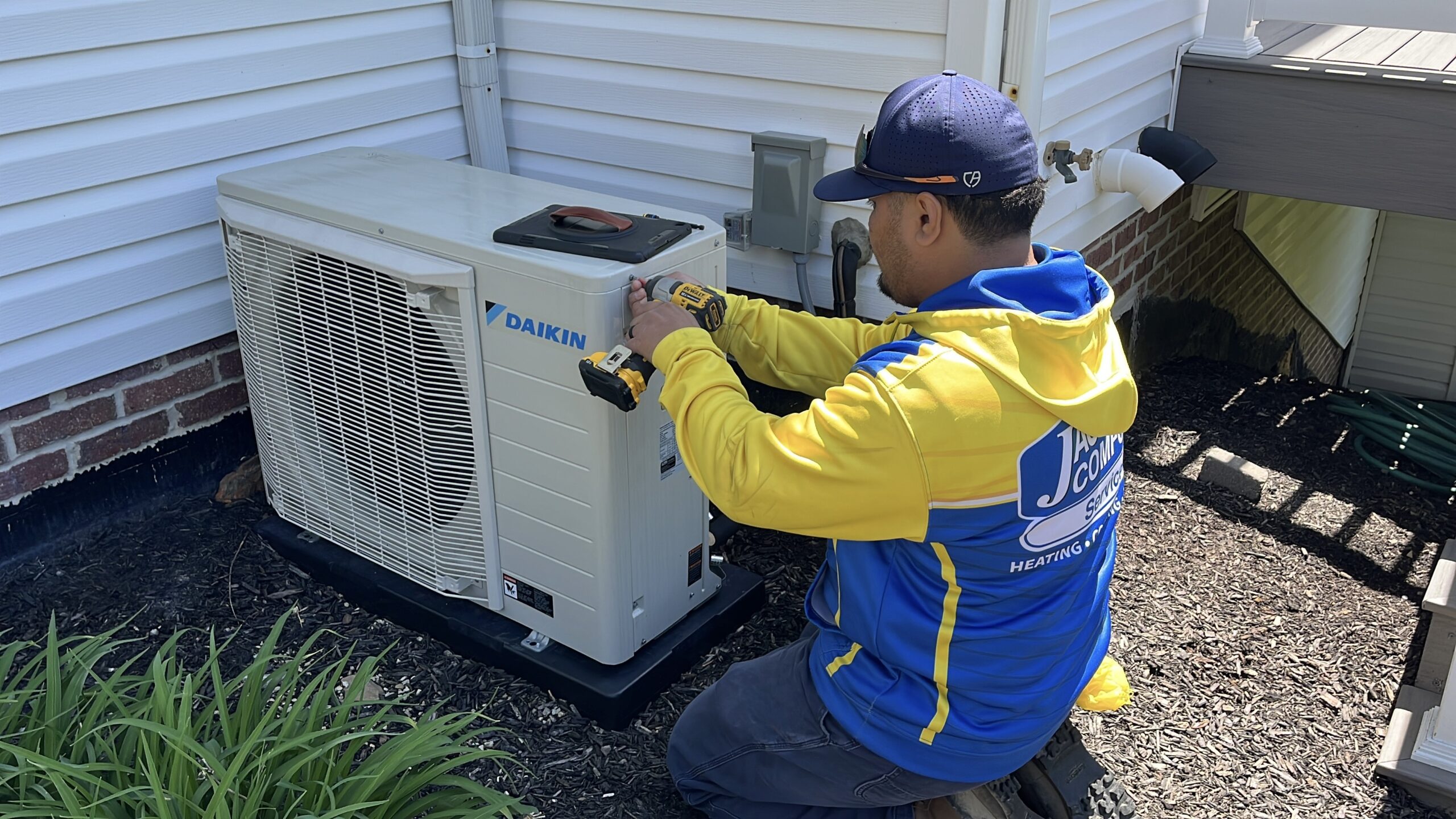 A technician in a blue and yellow uniform uses a power drill to work on an outdoor Daikin air conditioning unit next to a house.