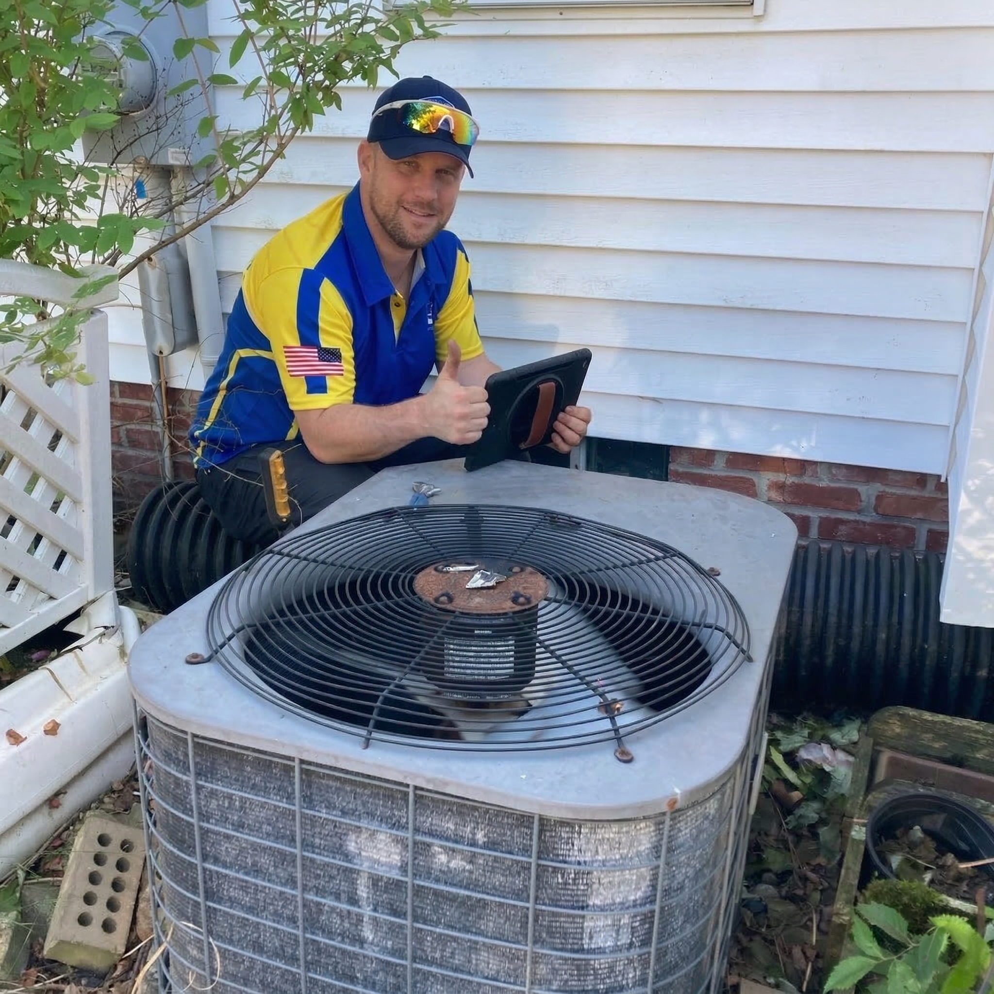 A Technician In A Blue And Yellow Uniform Kneels Next To An Outdoor Air Conditioning Unit, Holding A Tablet And Giving A Thumbs Up.